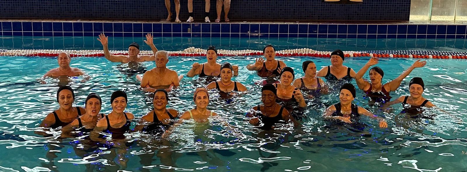 Turma de hidroginástica da 349 Fitness Experience dentro da piscina coberta. Cerca de vinte alunos da melhor idade, sorrindo, alguns com as mãos levantadas.